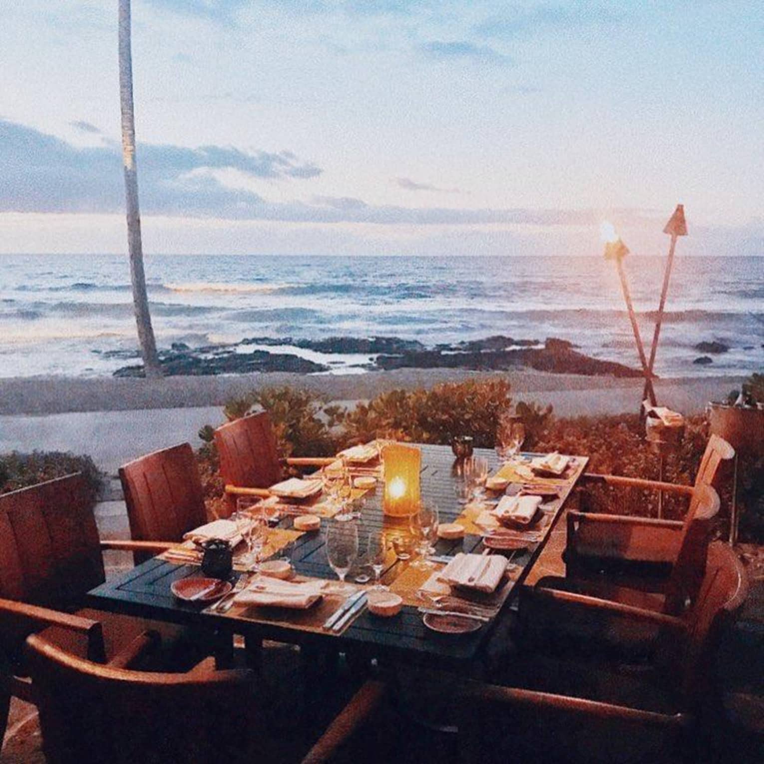A wooden dining table is set with dishes and lit candles by the beach at sunset