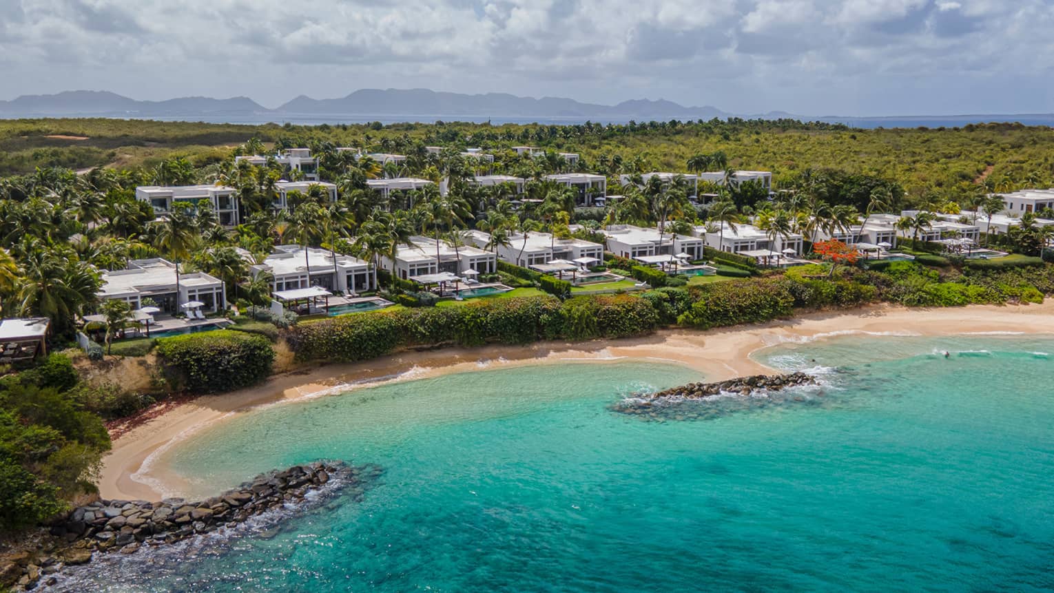 Structures among the trees on a verdant shoreline with a sandy beach and turquoise waters.