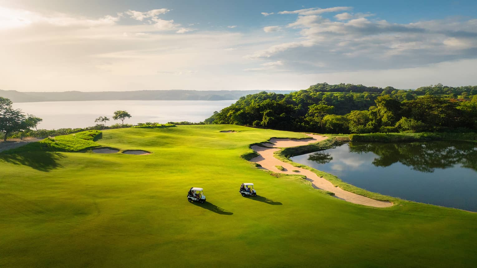 Two golf carts are parked on a fairway next to a lagoon with the ocean and tree-lined coastline in the distance
