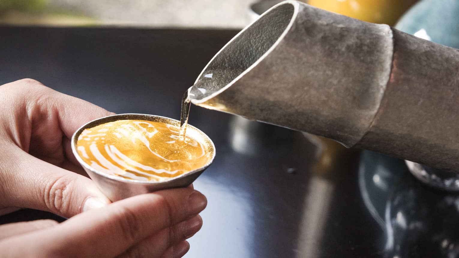Clear liquid taking on amber colour as it is poured from a hammered metal spout into small cone-shaped cup held by two hands