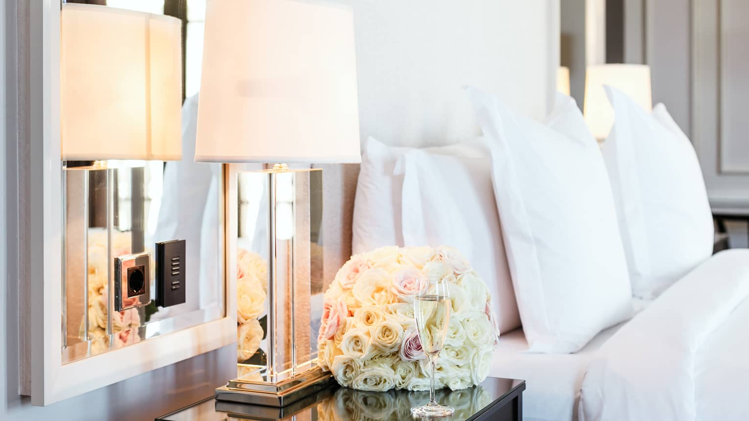 Detail of hotel room showing a spherical arrangement of roses and a flute of champagne on a nightstand with bed in background