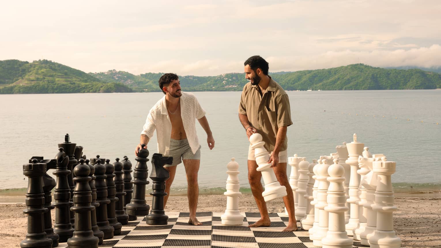 Two people wearing shorts and button-down shirts play giant chess on a beach