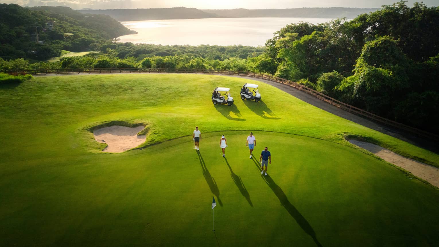 Aerial view of four people walking toward a green on a golf course with two golf carts parked nearby and the ocean in the distance