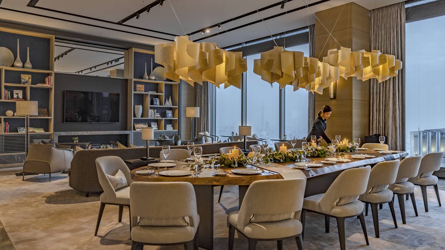 A candlelight dinner is set at a long, oval table with plush seating for twelve under pendant lamps made of swirling wood.