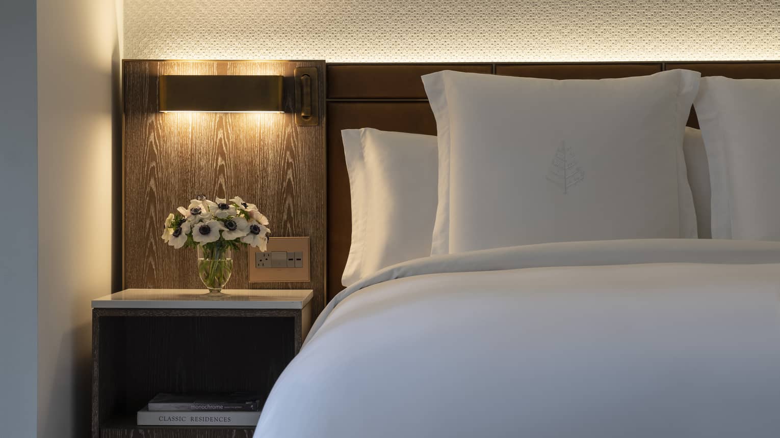 Close-up of bed with white linens and pillows, light above bedside table with vase of flowers