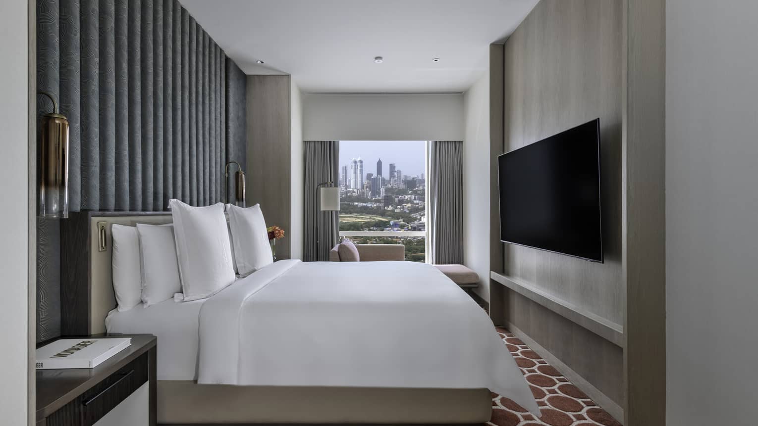 Renovated Executive Suite bedroom with bed and white linens, salmon chaise lounge next to floor-to-ceiling window, grey accent wall, wall-mounted TV, red patterned carpet