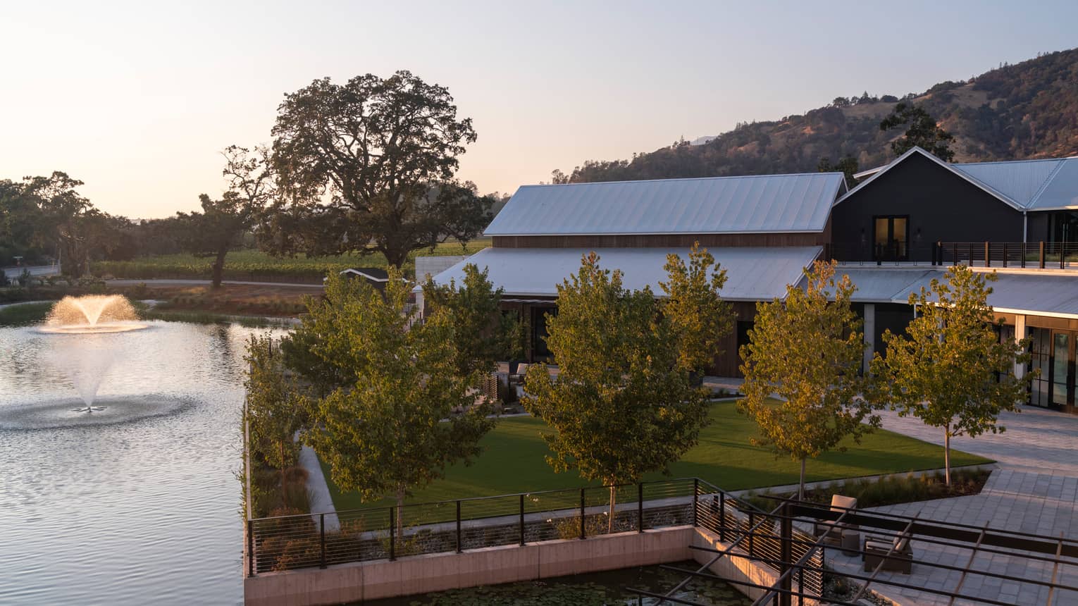 A spacious green lawn space surrounded by trees, a seating area and a lake with a fountain