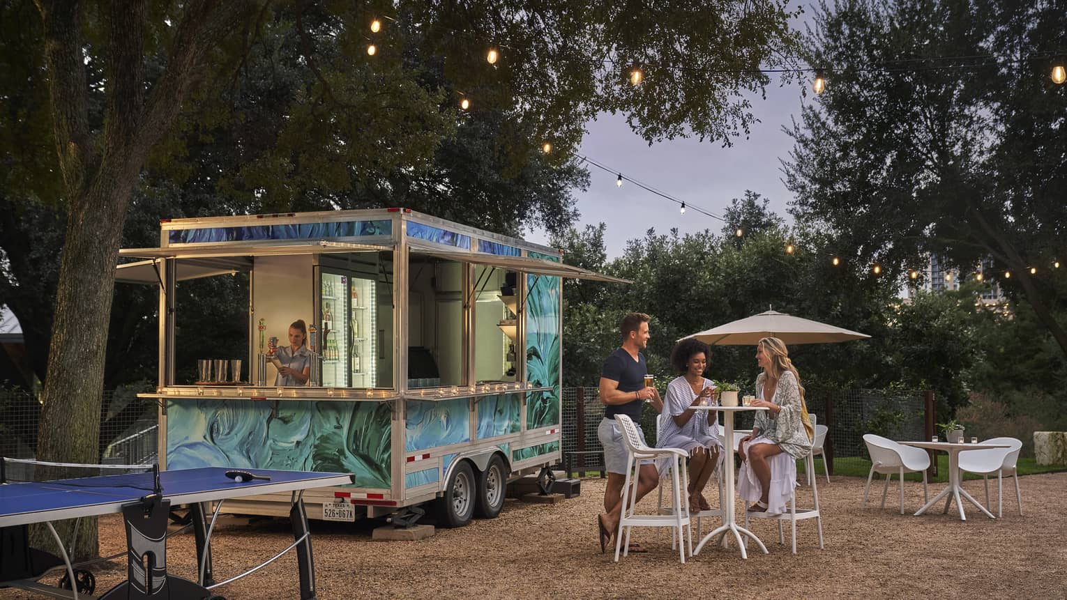 Three people sitting outside of a food truck surrounded by trees.