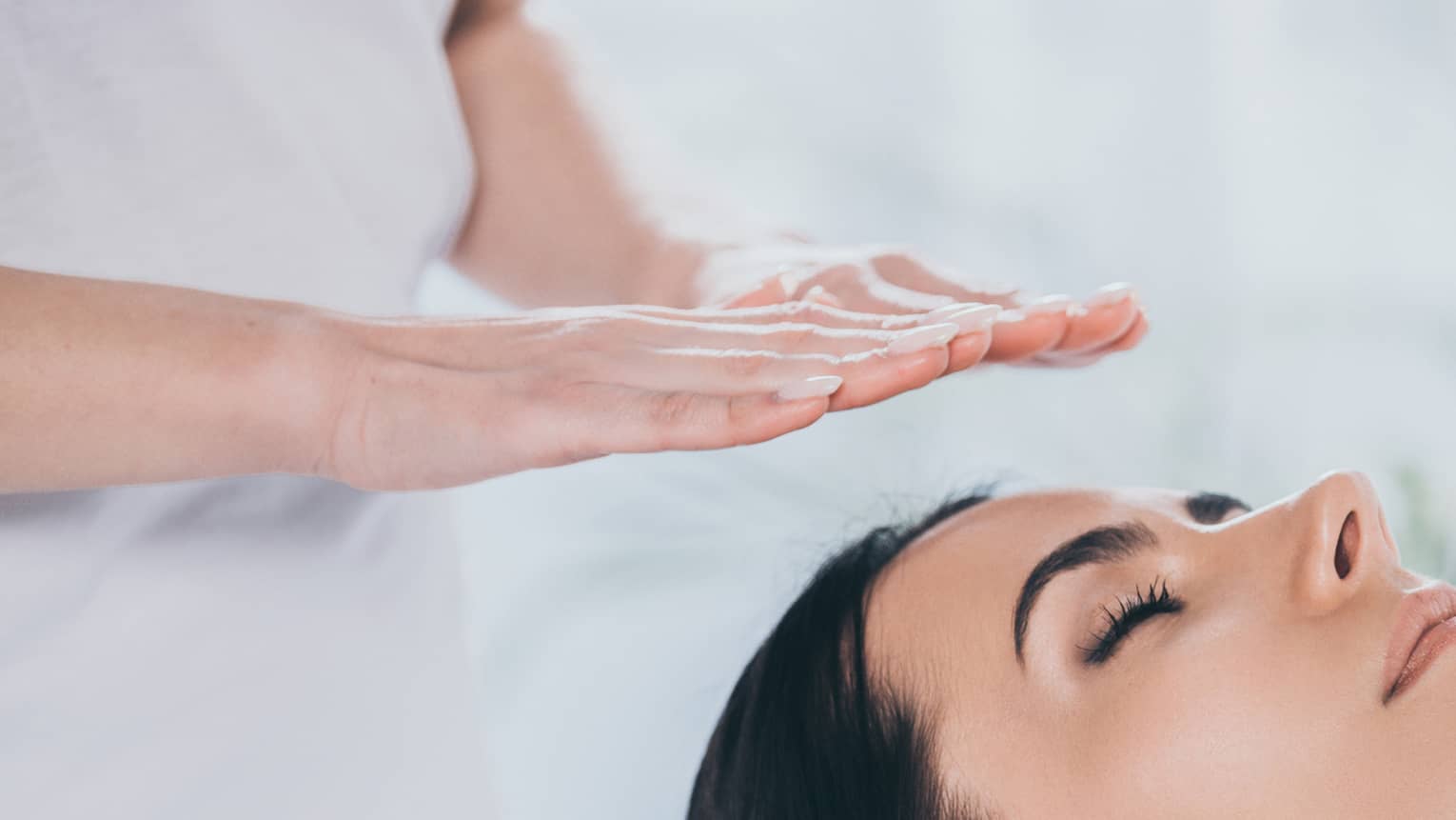 Woman with eyes closed lays on rolled up towel on spa table under therapists' hovering hands
