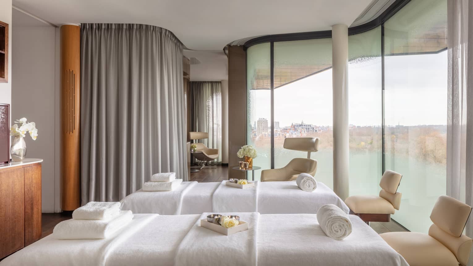Spa couples treatment room with two white beds, chairs at ends, tall windows with city views