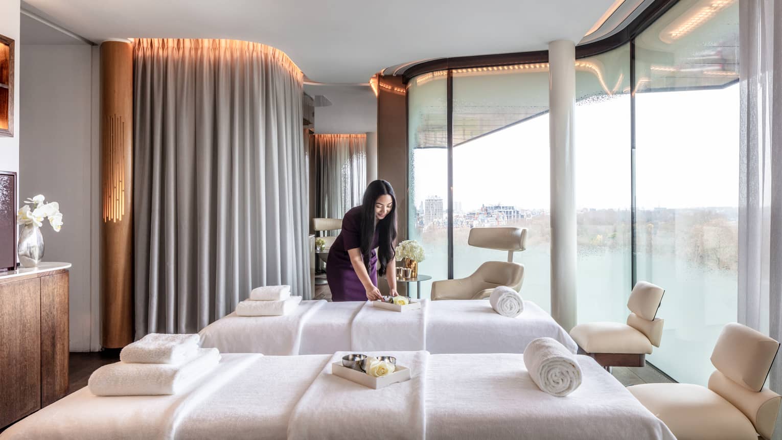 Spa couples treatment room with staff member, two white beds and chairs at ends, tall windows with city views