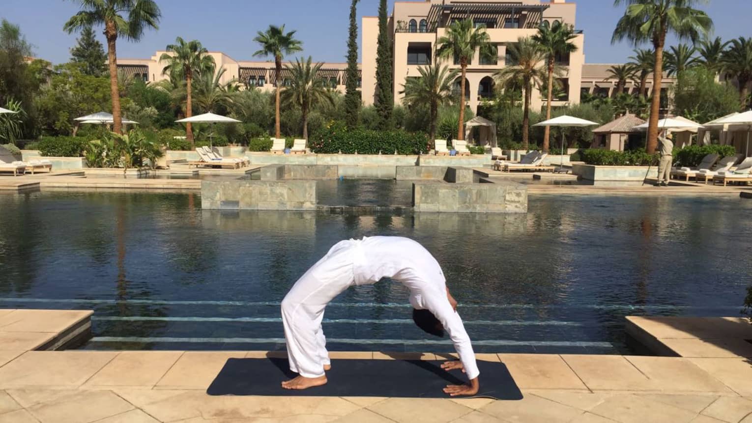 Person wearing white poses in arched yoga bridge pose beside outdoor swimming pool
