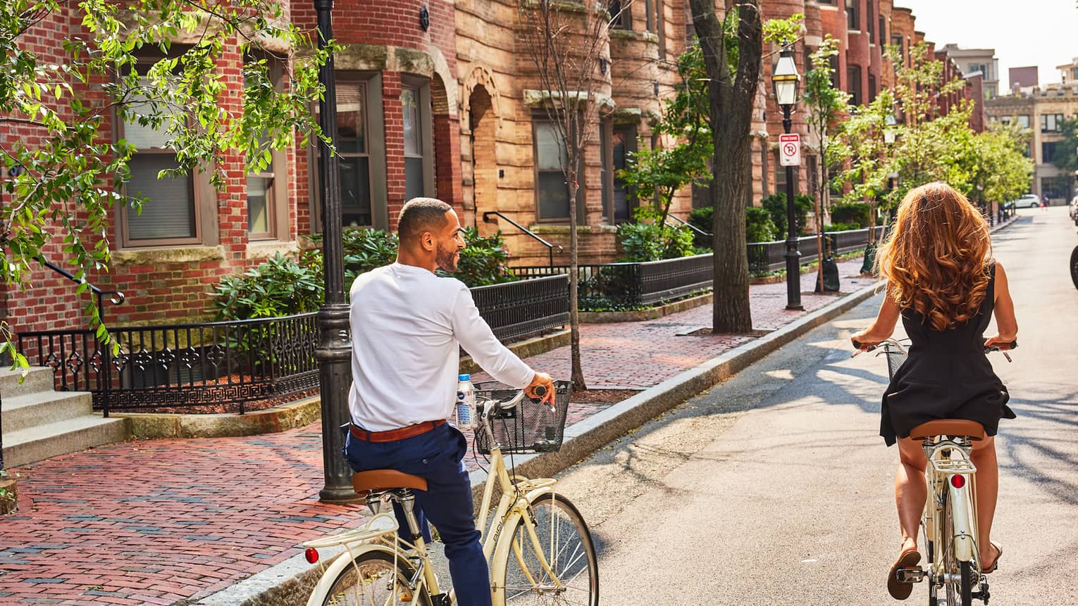 A couple rides bicycles along a street lined with brownstone homes