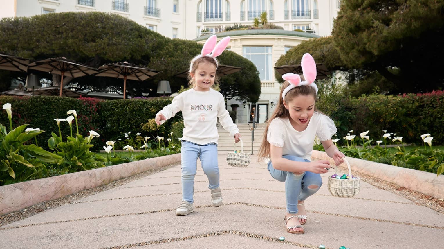 Two children in matching outfits wearing pink rabbit ears and holding Easter baskets with grand white building in backdrop