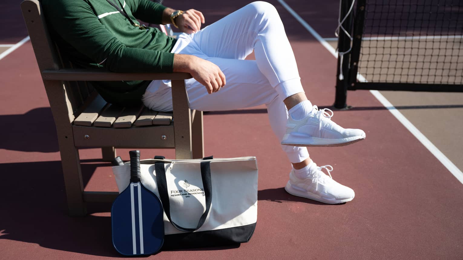 A person sits on a wood bench on the sideline of a pickleball court. A tote bag and pickleball racquet are next to the bench.