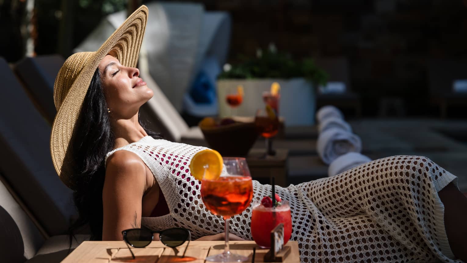 A guest laying in a lounge chair in the sunshine with their sunglasses and drink on a table beside them
