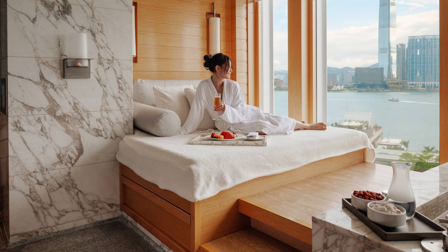 Woman in spa bathrobe sits on windowside chaise looking out to Victoria Harbour at Four Seasons Hotel Hong Kong