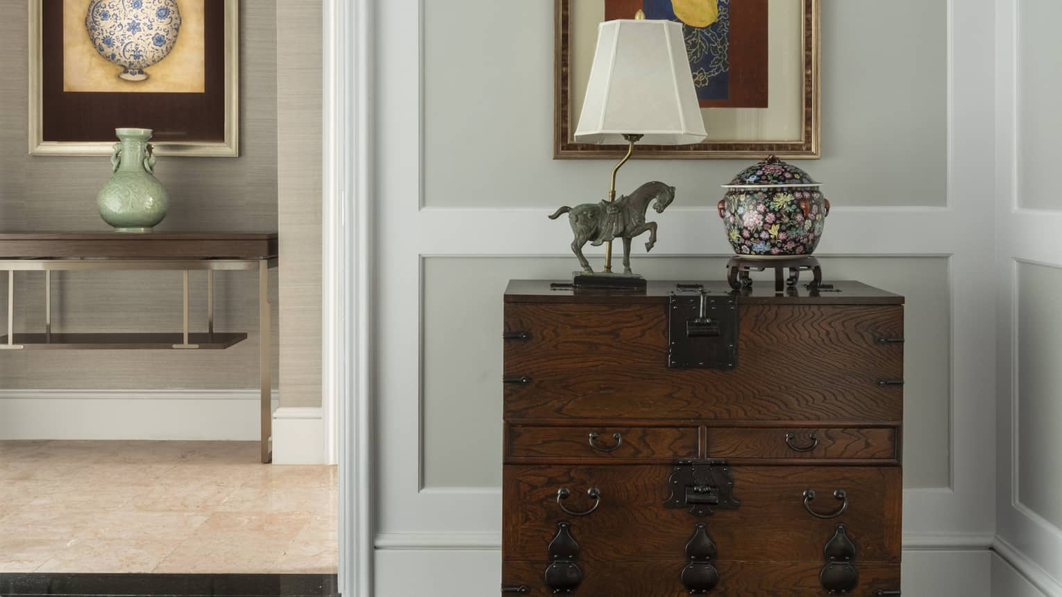 A decorative hallway with a wooden chest adorned with a table lamp, horse statue and vase. Framed artwork hangs above, with a view of another room displaying a vase and painting.