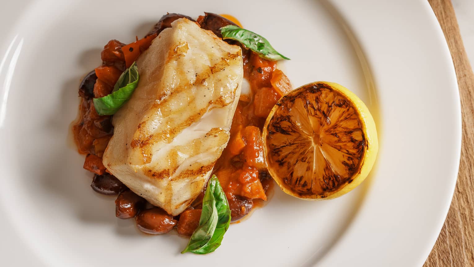 Piece of grilled white fish on bed of stewed vegetables, grilled half lemon and basil garnish on round white plate