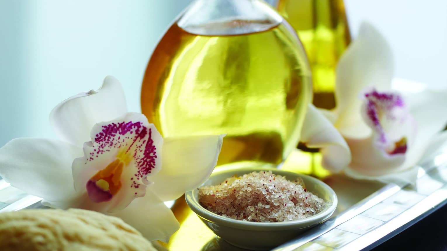 Close-up of small spa bowl with salt scrub, glass bottle with oil, white and purple tropical flower