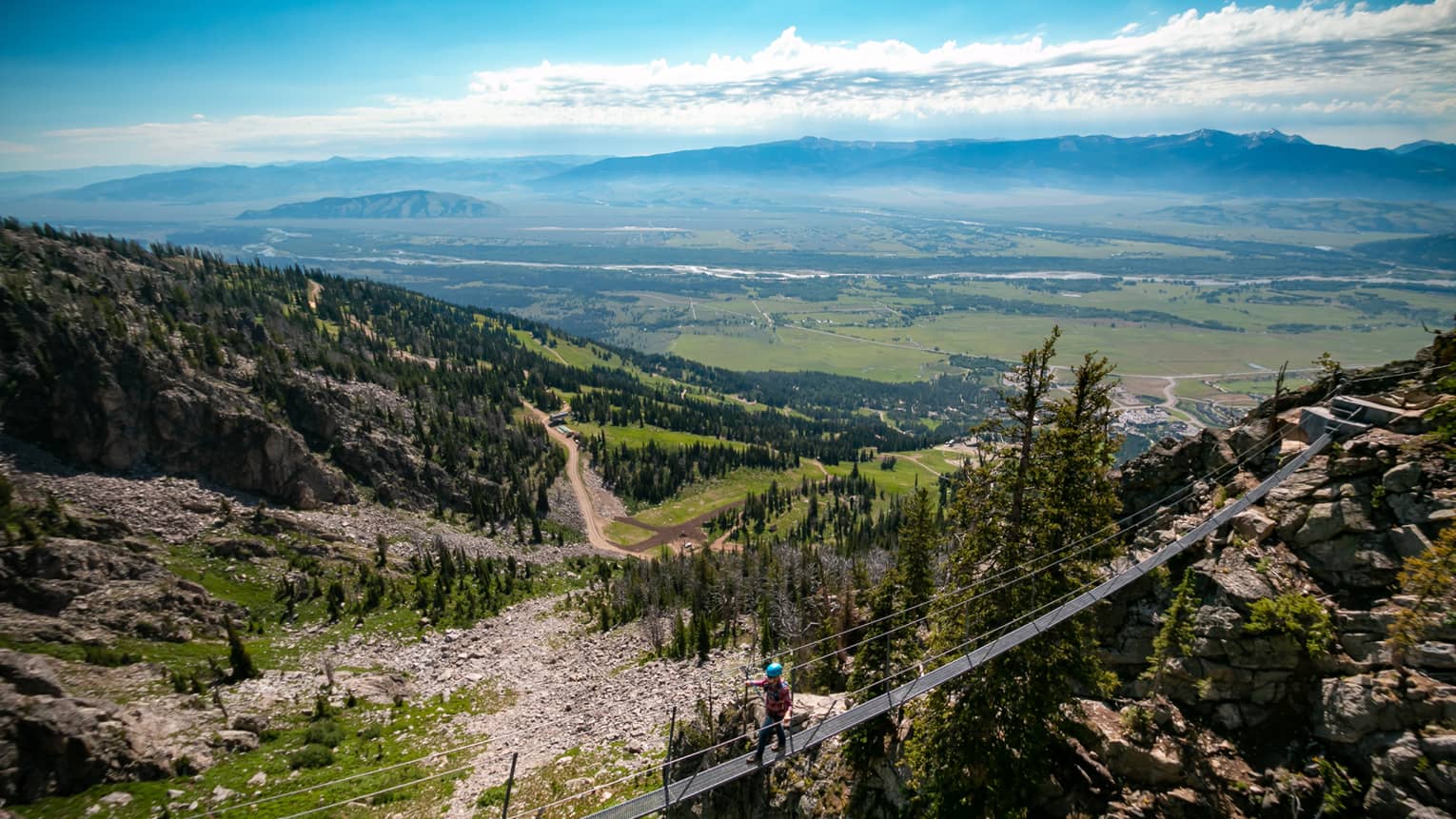 A man walks across a tightrope, a view of the lush green landscape below him 