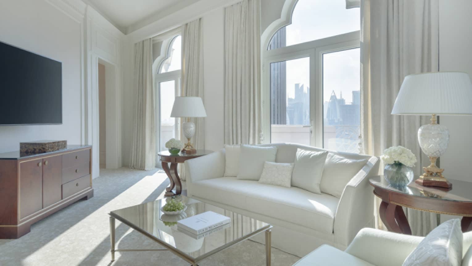 A light-filled living room with white sofa, white arm chair, tan carpet, glass coffee table, lamps