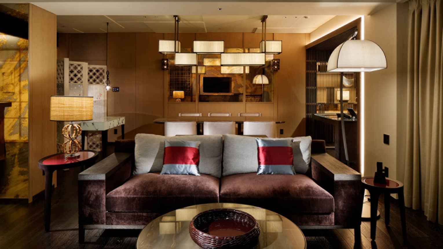 Living room with dark plum?coloured sofa with two silver accent pillows. Behind the sofa is a dining room illuminated with multi-shade chandelier.