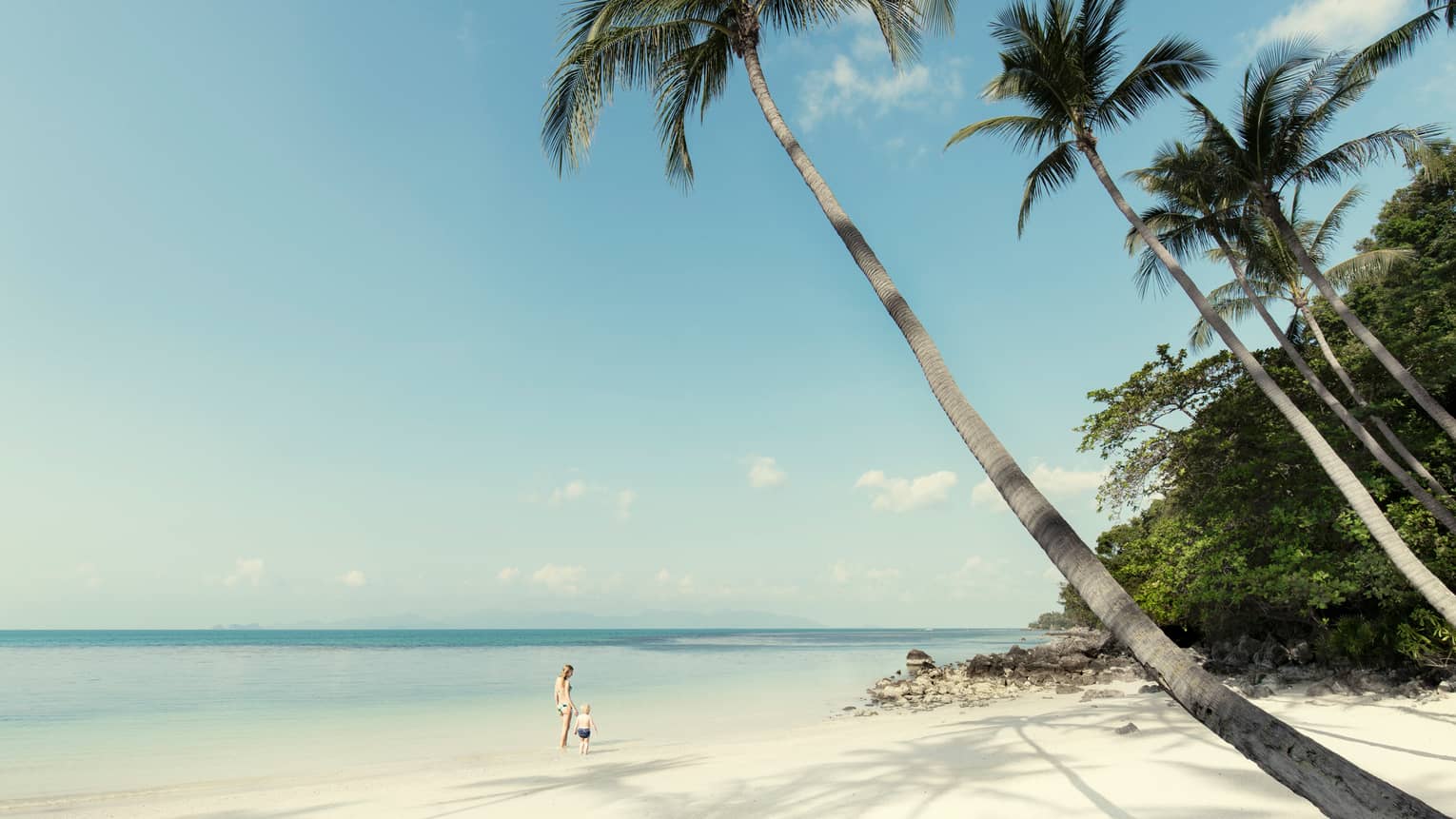 Mother and toddler wearing swimsuits walk along white sand beach, tide under clear blue sky