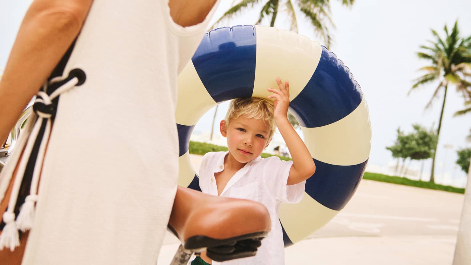 Young child wearing green swim trunks and a white shirt holds a blue-and-cream pool float while walking next to adult wearing a cream-coloured beach cover up and walking next to a bicycle
