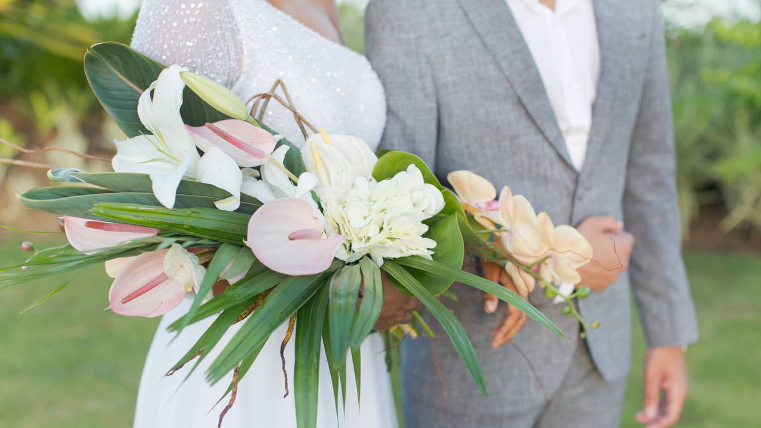 Groom and bride holding large tropical wedding bouquet walk across lawn