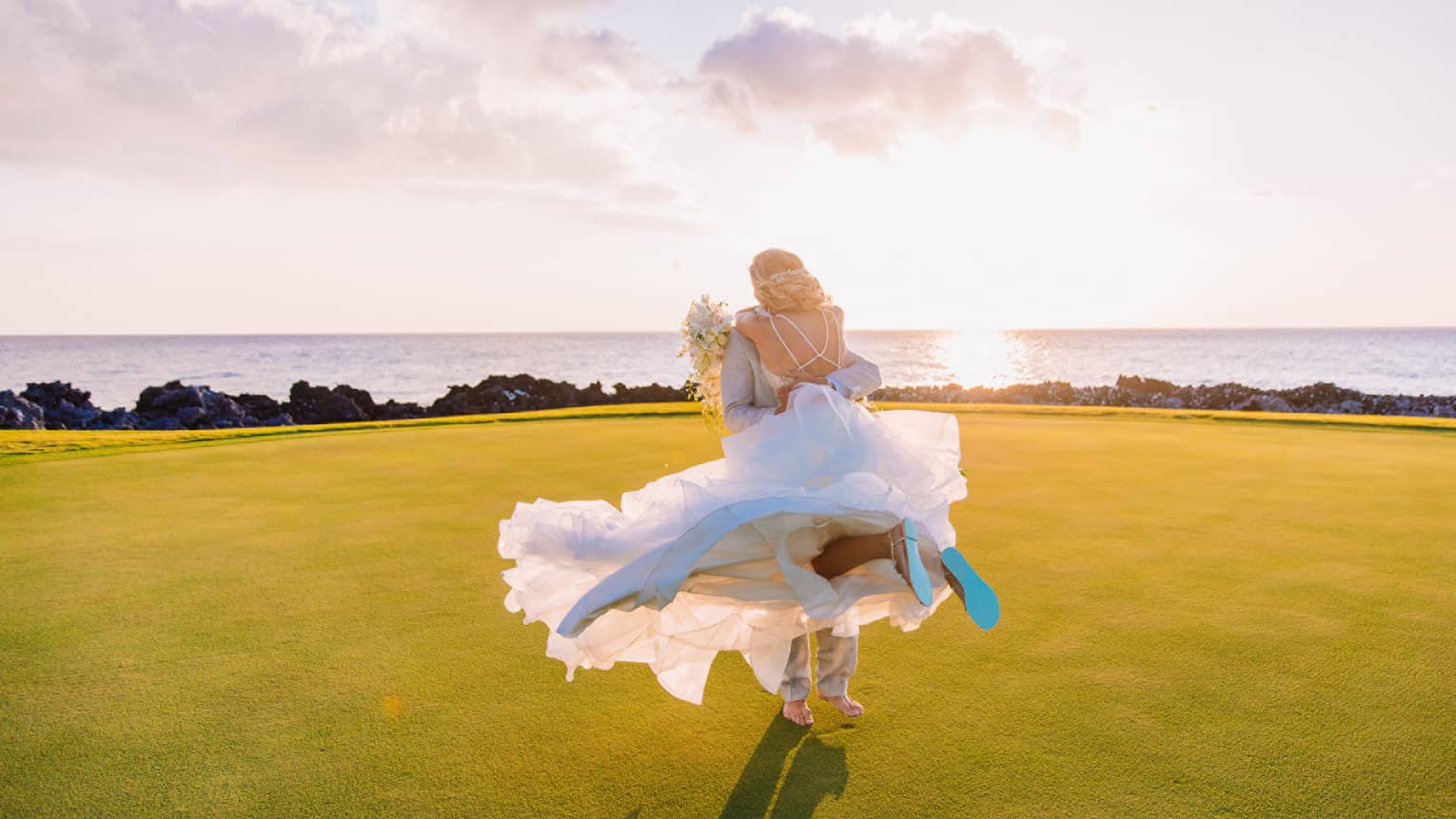 Groom lifting bride off ground on green lawn near ocean