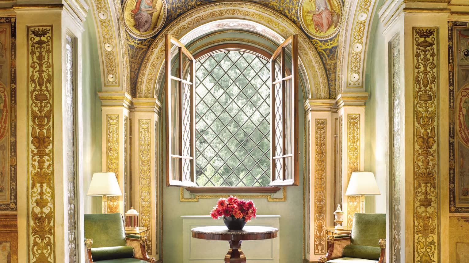 Green velvet armchairs around wood table with flowers, open window, gold ornate pillars