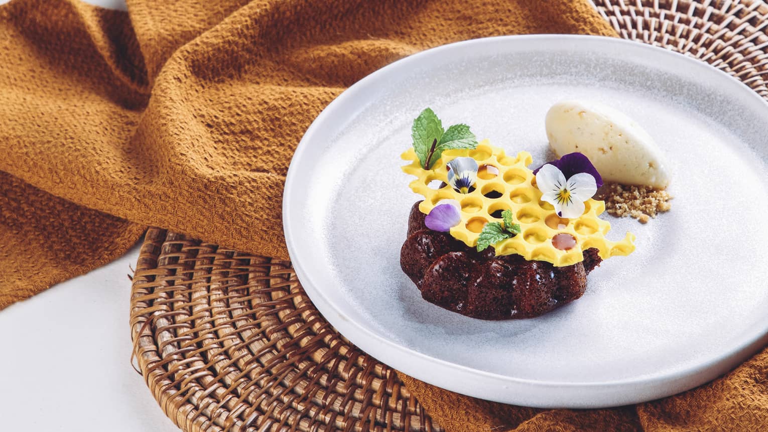 A rich chocolate mini-cake topped with honeycomb-shaped white chocolate garnished with mint leaves and dainty flower petals