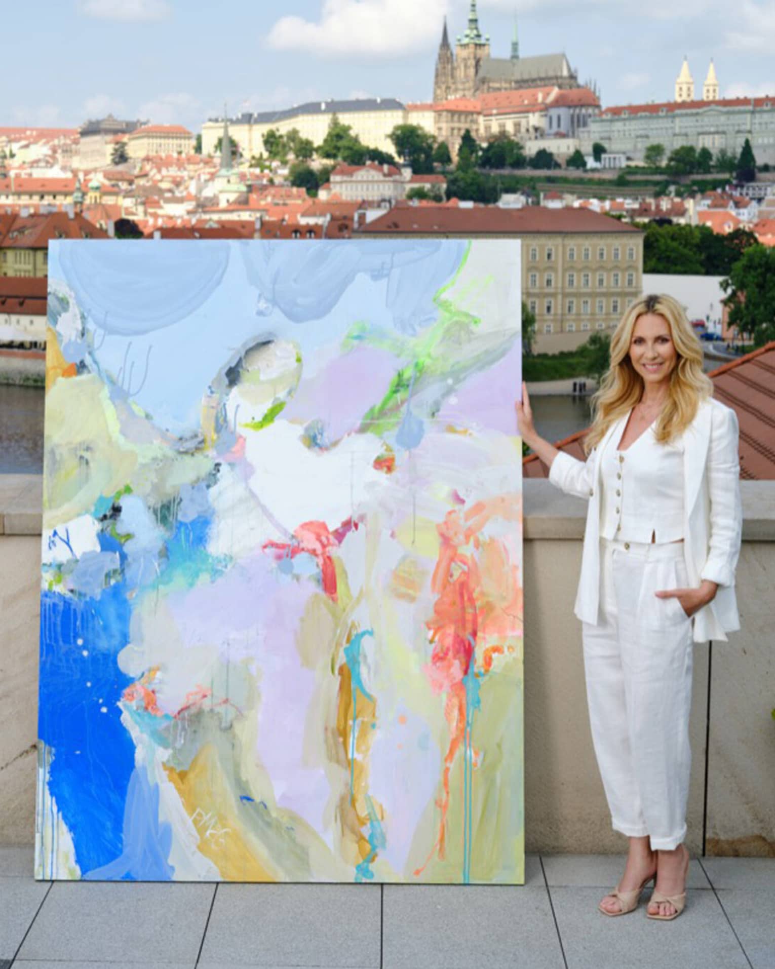 Artist Petra McGrath stands next to a colorful painting on a rooftop overlooking the city of Prague