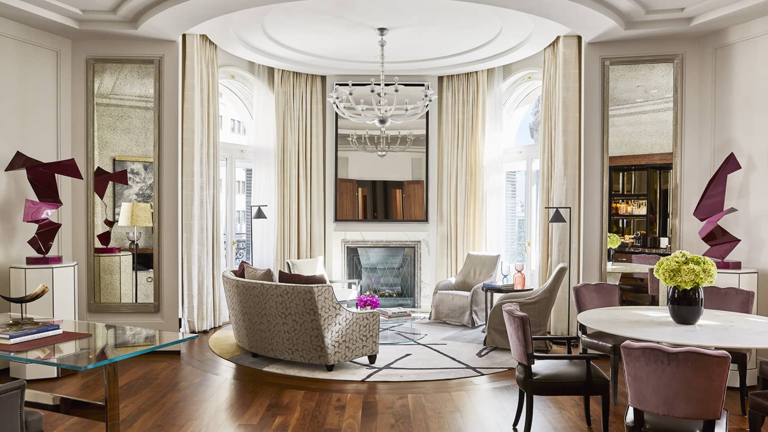 Luxury hotel suite with round dining table, sculptural decor, desk, curved sofa, chairs, fireplace, large mirrors, chandelier and arched windows with drapes