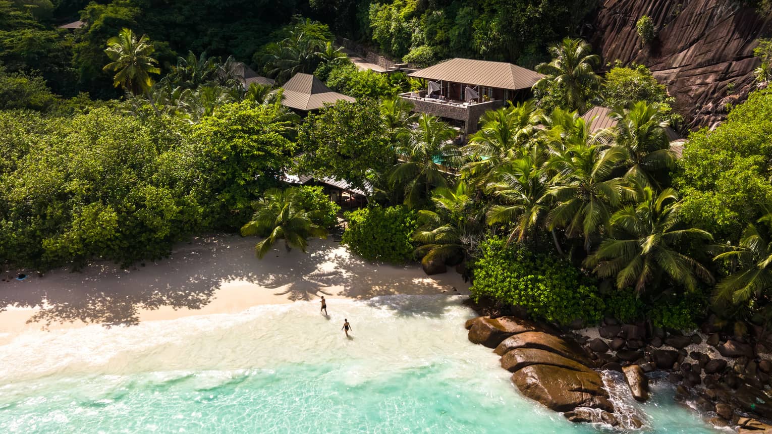Aerial view of a secluded resort villas nestled in a lush green landscape near a white-sand beach with clear turquoise waters