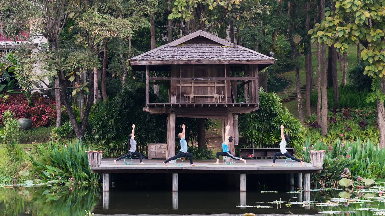 Four people doing yoga on a deck overlooking a calm pond, in front of a wooden bungalow and jungle trees