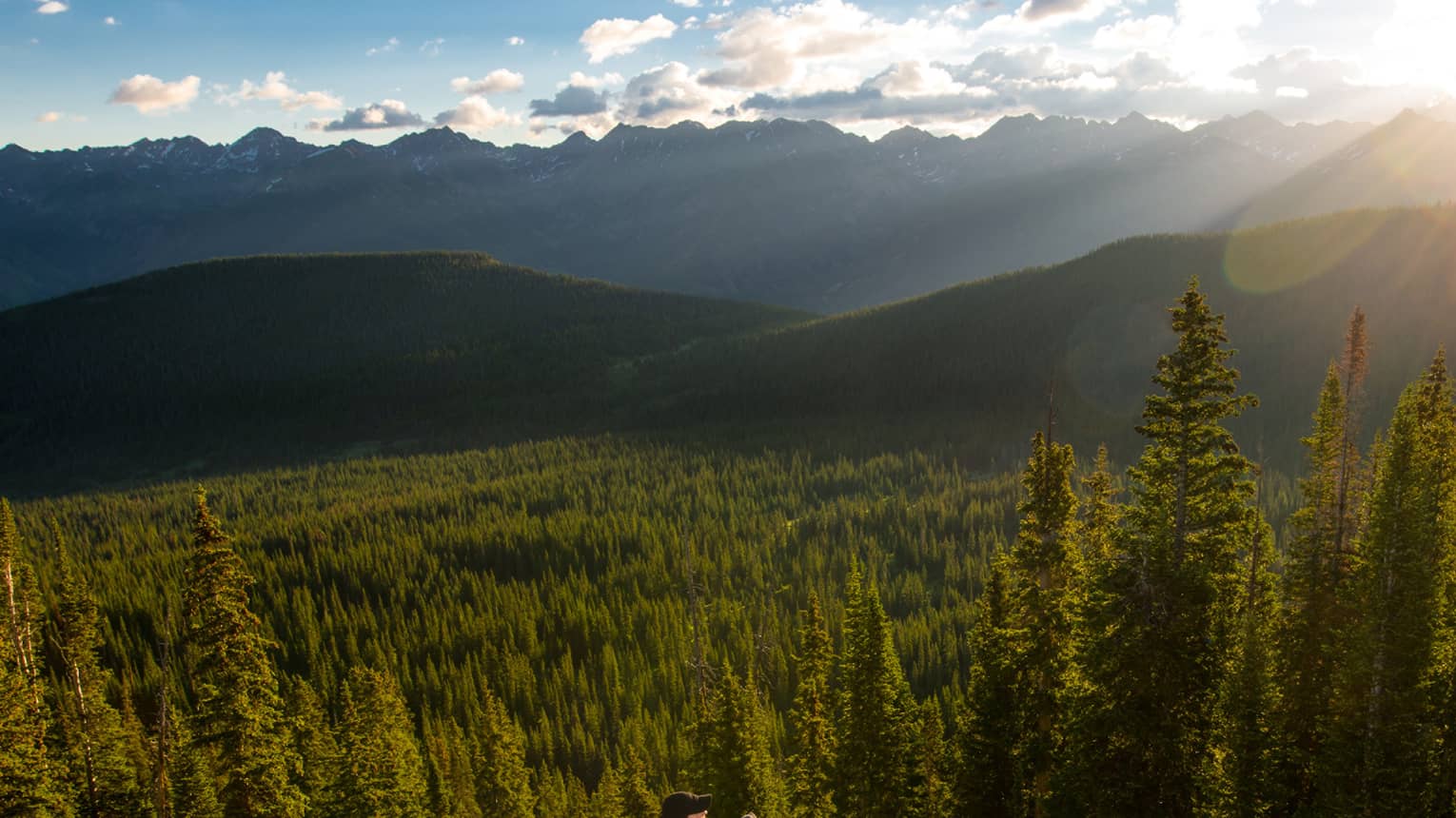 Person sits at edge of cliff overlooking trees, mountains as sun starts to set