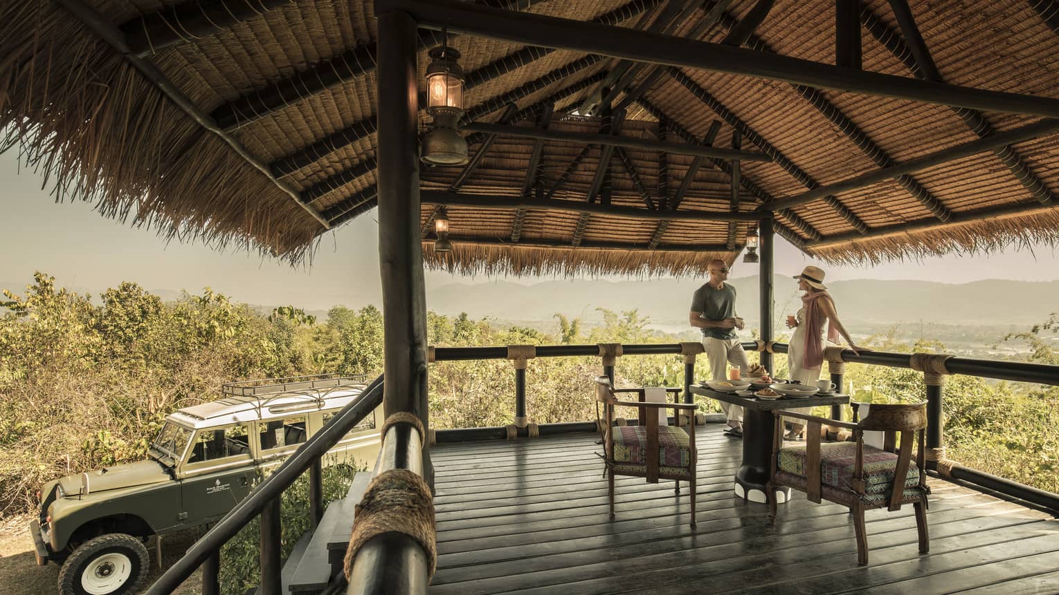 A couple sitting on a wooden deck overlooking green brush, Land Rover sitting nearby