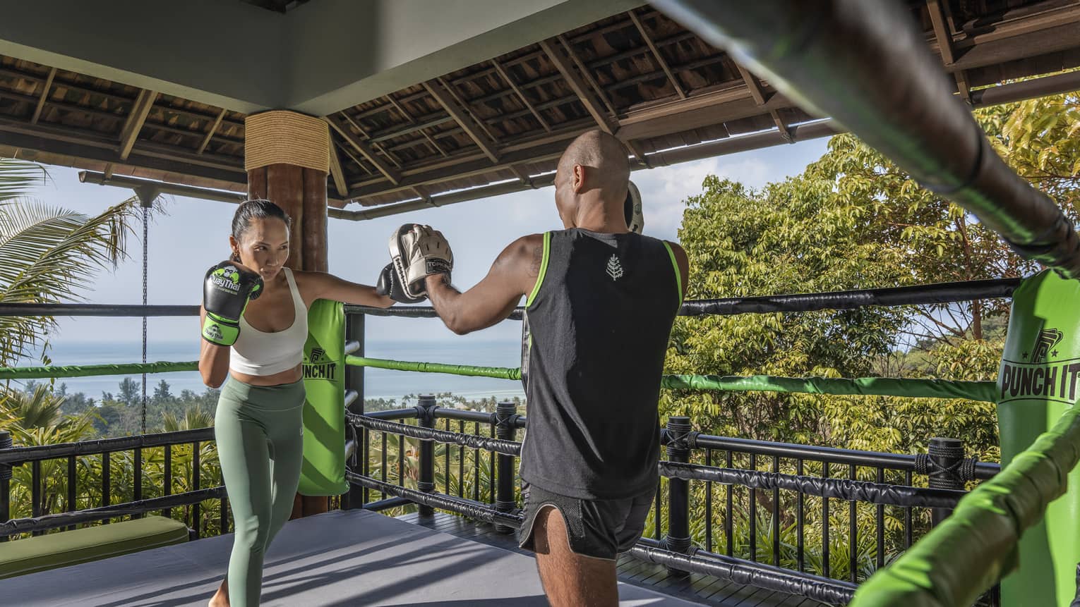 Two people practise Muay Thai in an outdoor boxing ring nestled in the treetops and overlooking the forest and the ocean.