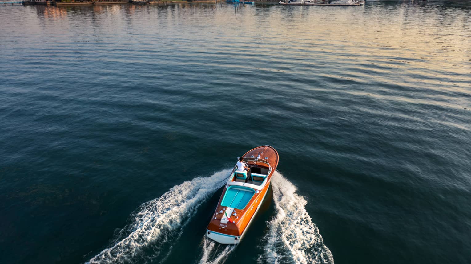 Person driving a wooden speedboat across a lake towards shore at sunset