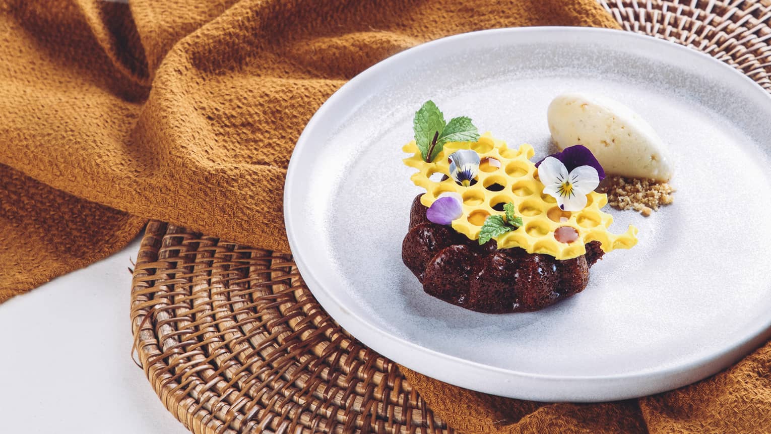A rich chocolate mini-cake topped with honeycomb-shaped white chocolate garnished with mint leaves and dainty flower petals