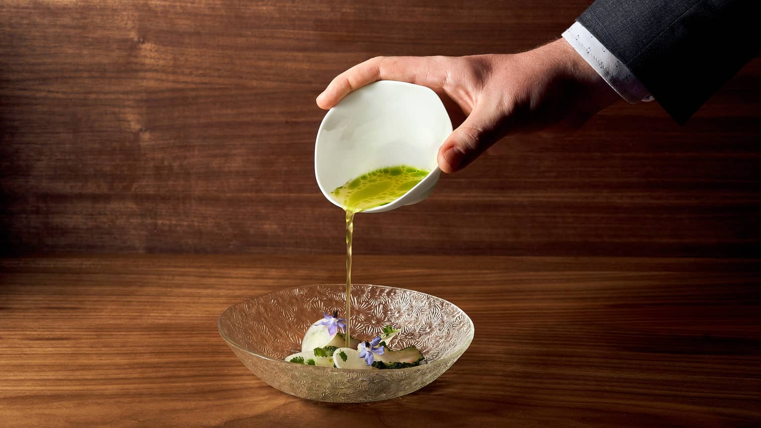 A close-up shot shows a hand delicately pouring a vibrant green sauce from a white bowl onto an intricately designed glass plate.