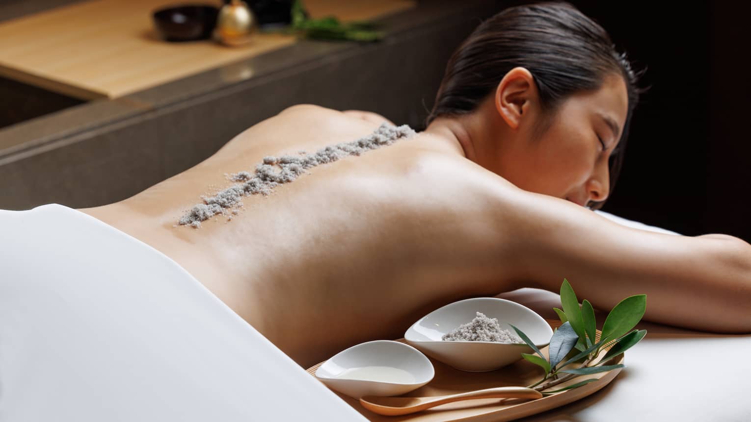 Female guest lies facedown on spa treatment table with line of exfoliating sand running the length of her spine