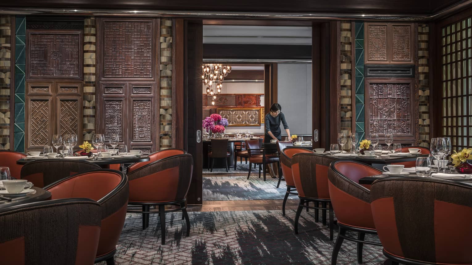 Woman sets table at Jiang-Nan Chun, Michelin-starred Cantonese restaurant, with red leather bucket chairs around dining tables, carved wood walls.