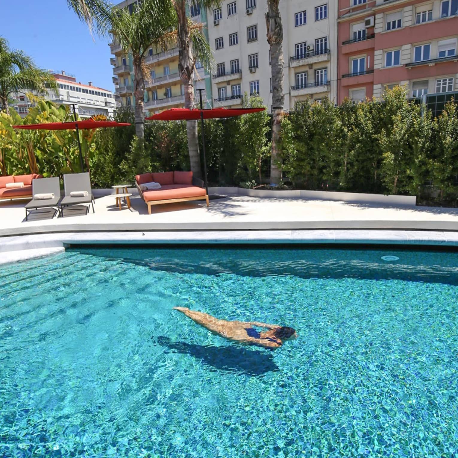 Person swimming in a clear outdoor pool surrounded by palm trees, lounge chairs and colourful apartment buildings on a sunny day.