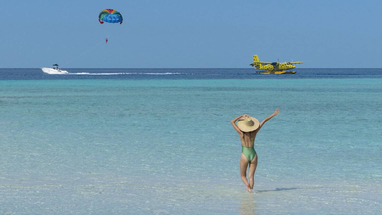 A woman walking on the beach watches a seaplane and a parasailor in the distance.