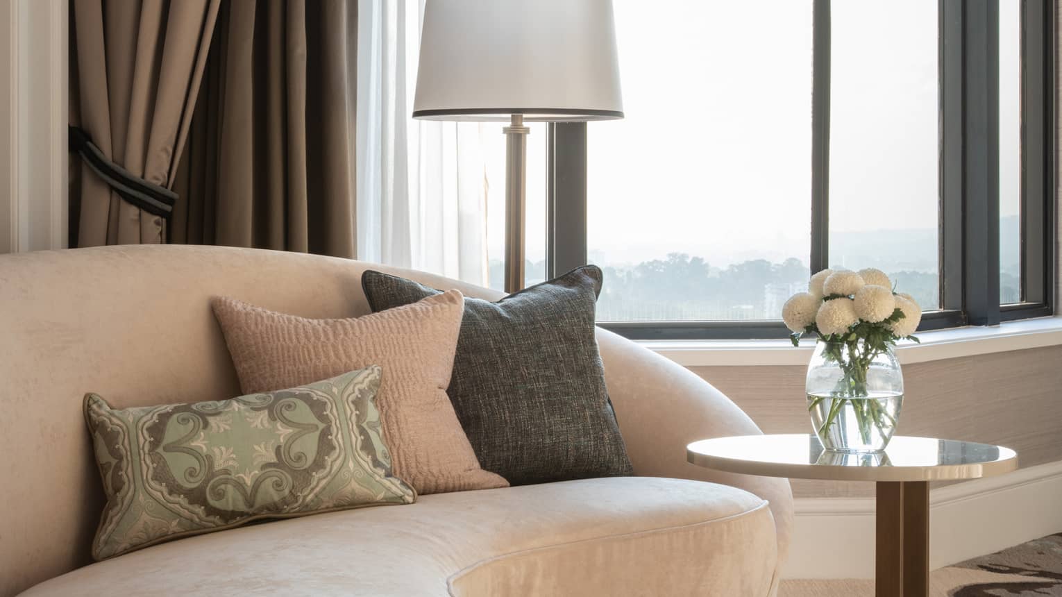 A cosy seating area with a beige curved sofa, decorative pillows, side table with white flowers and floor-to-ceiling windows offering natural light and outdoor views.