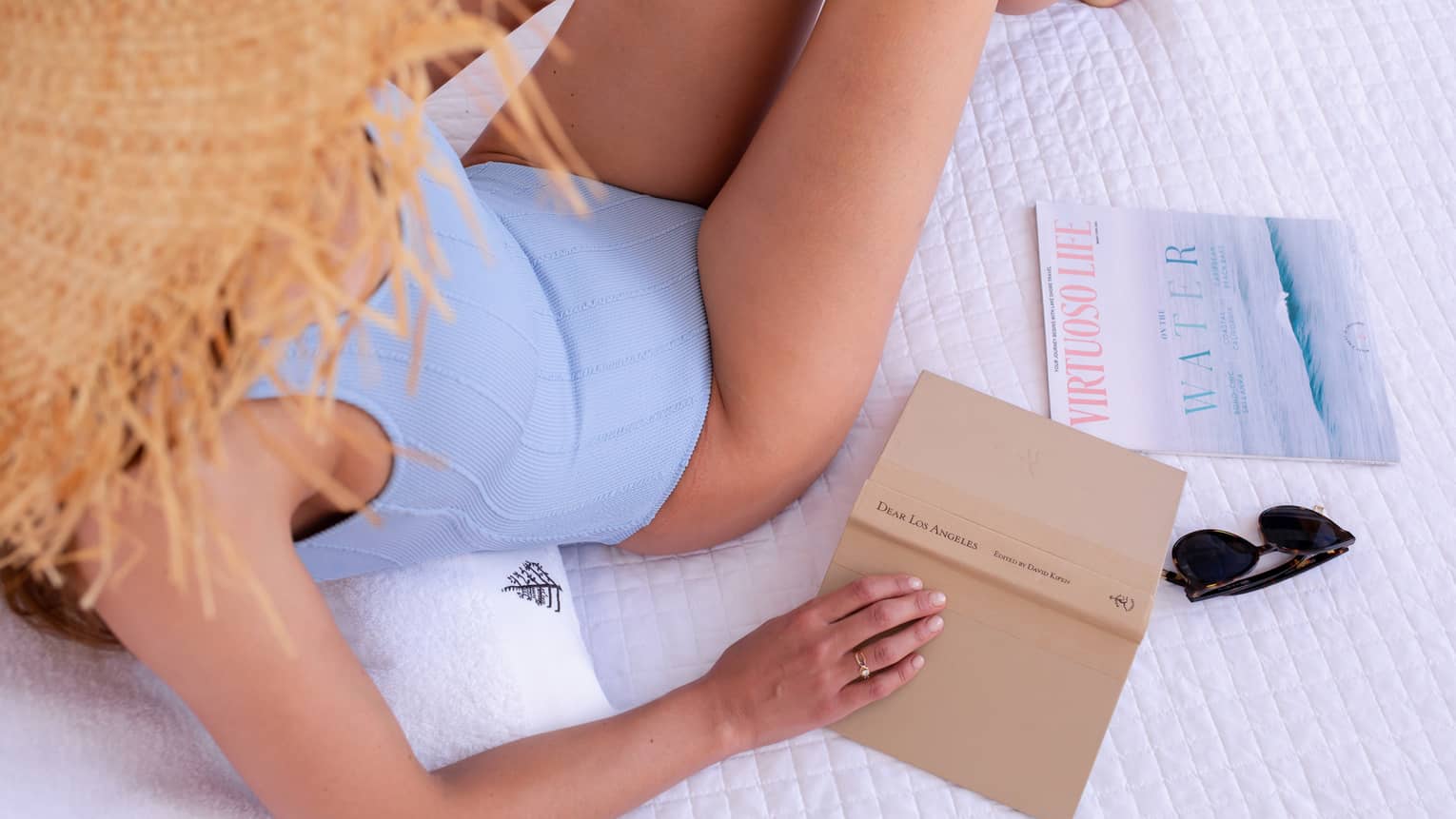 A woman in a blue swimsuit and straw hat holding a tan book while lounging on white bed