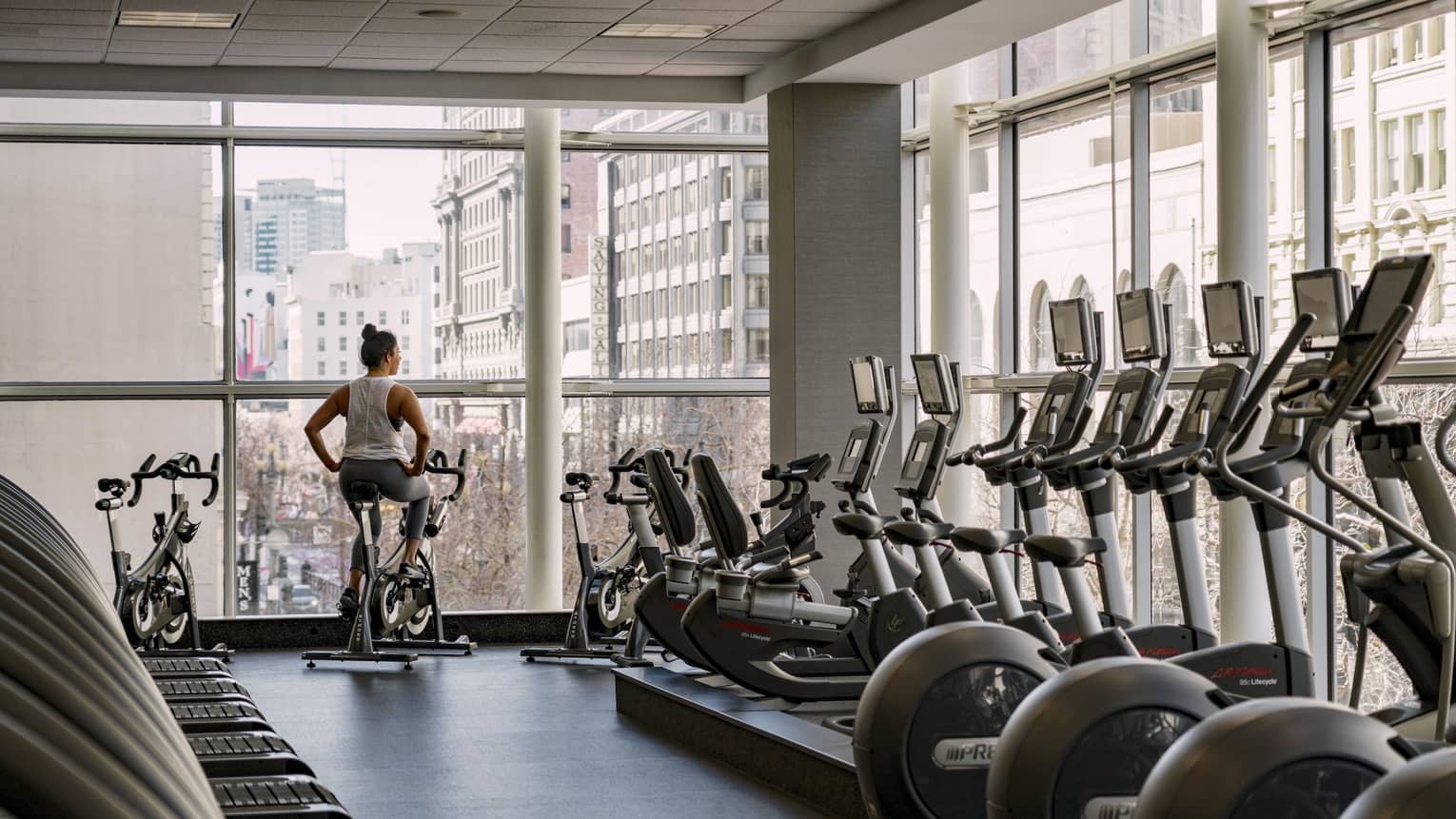 A woman rides a stationary bike in a hotel fitness center with floor-to-ceiling windows overlooking a city view